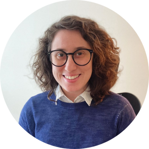 A person with curly hair and glasses smiles at the camera, wearing a white shirt under a blue sweater.
