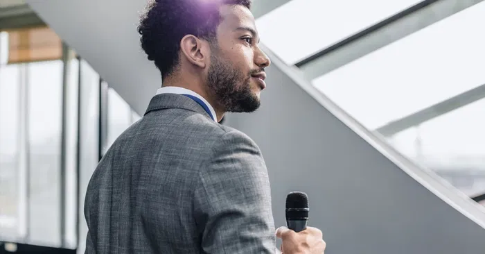 Person in a suit holding a microphone, speaking indoors with large windows in the background.