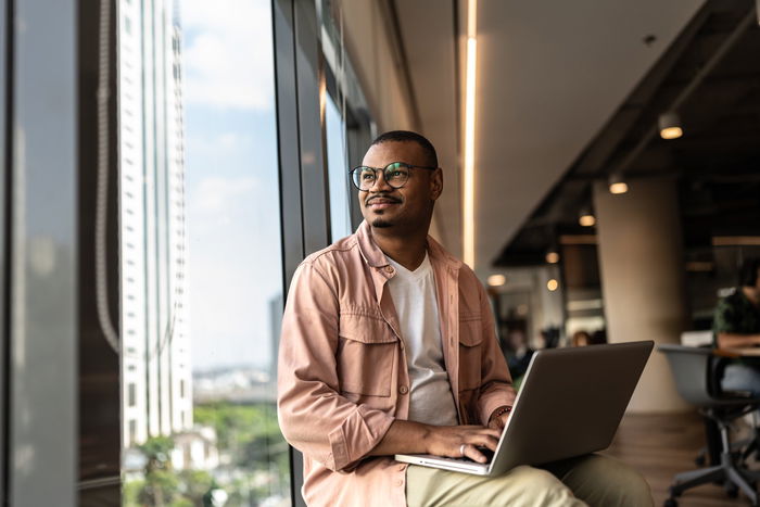 A person sits by a large window in a modern office, holding a laptop and looking outside with a neutral expression.