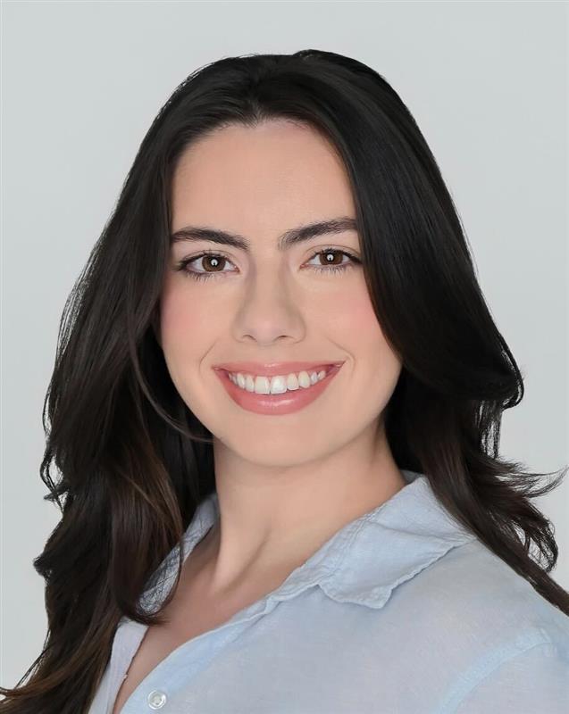 A person with long dark hair smiling, wearing a light blue shirt, photographed against a plain background.