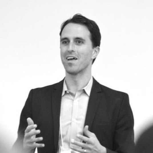 A man in a suit speaks, gesturing with his hands, in a black and white photo.