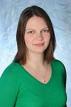 A woman with shoulder-length brown hair wearing a green top against a textured light blue background.