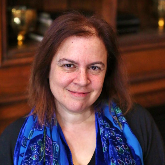 A person with shoulder-length brown hair wearing a blue patterned scarf smiles at the camera.