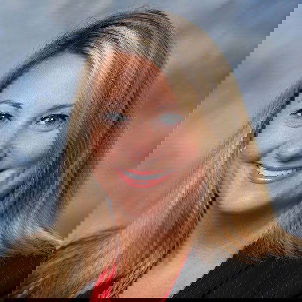 A person with long blonde hair and a black blazer smiles against a soft blue background.