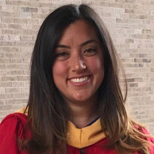 A person wearing a graduation gown smiles in front of a brick wall.