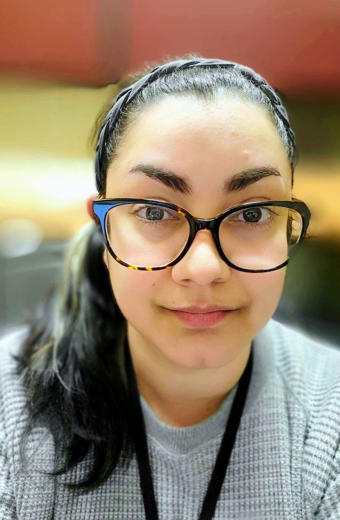 A person with glasses and a headband looks at the camera in a well-lit room, wearing a gray sweater.