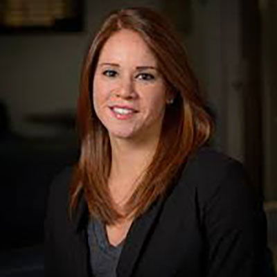A woman with long, straight brown hair and a black blazer smiles at the camera in an indoor setting.