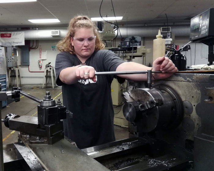 Person using a lathe machine in a workshop, wearing safety glasses and focusing on adjusting a component.