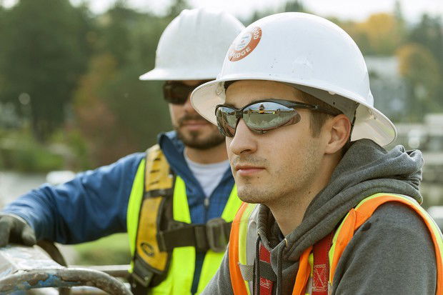 Two construction workers wearing helmets, reflective vests, and sunglasses stand outdoors at a work site.