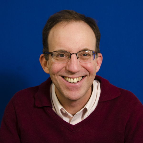 A person wearing glasses and a maroon sweater smiles at the camera against a blue background.