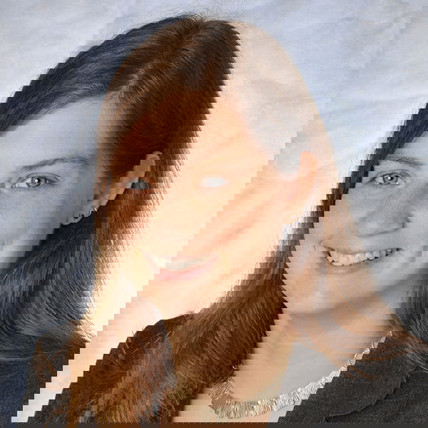 A woman with long brown hair, wearing a gray blazer and a necklace, smiles against a light background.