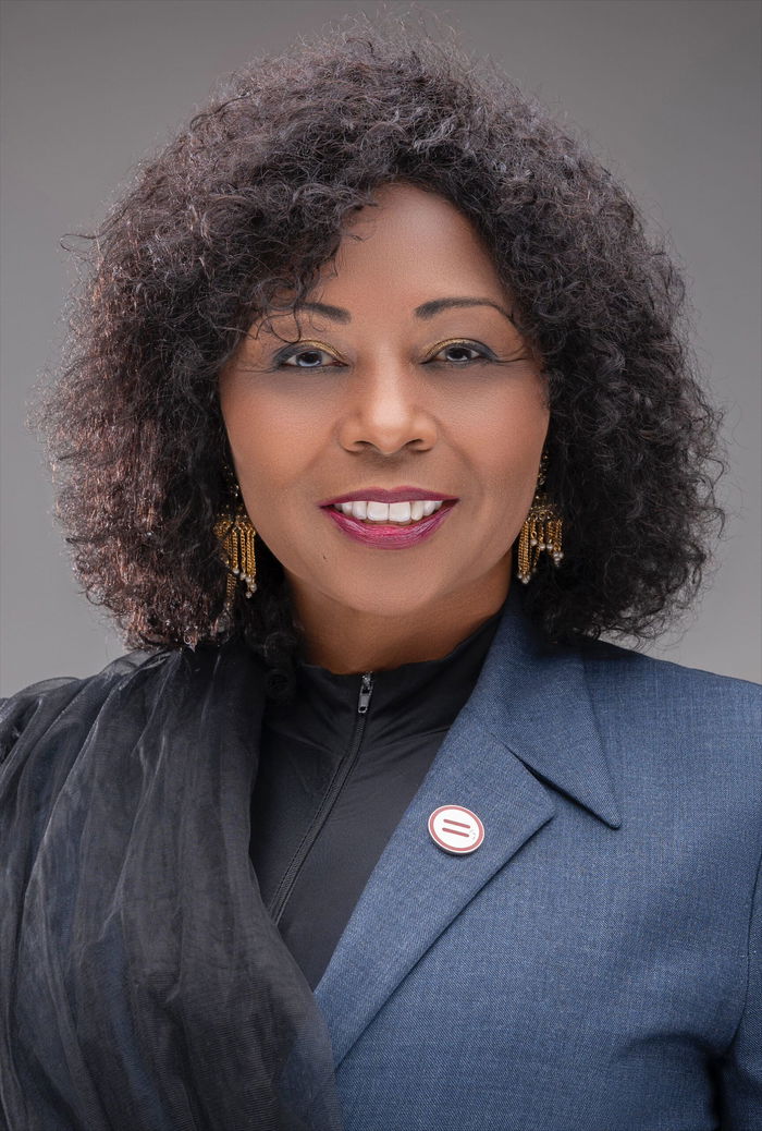 A person with curly hair wearing a blue blazer and a black top, smiles at the camera.