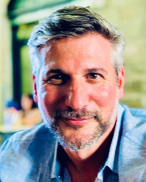 Man with short gray hair and a beard, smiling while wearing a blue shirt, with an out-of-focus background.