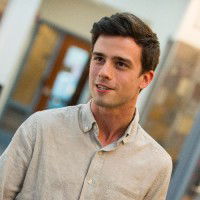 A person in a light button-up shirt stands indoors with a blurred background.