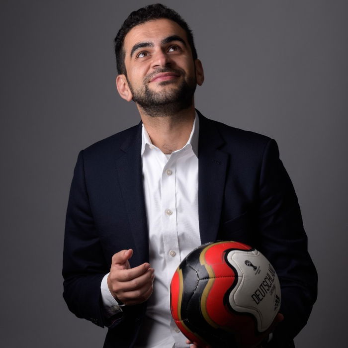 A man in a suit holds a soccer ball, looking upwards against a gray background.