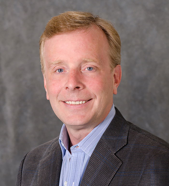 A man with short light brown hair wearing a dark blazer and blue shirt against a gray background.