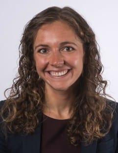 Woman with curly brown hair smiling against a plain background, wearing a dark blazer and top.