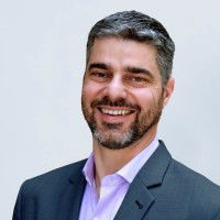 A man with a beard and short hair is smiling, wearing a dark suit jacket and a light purple shirt, against a plain background.