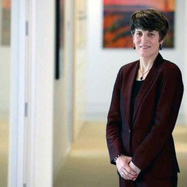 A person in a dark suit stands in a hallway with art on the walls, smiling slightly at the camera.