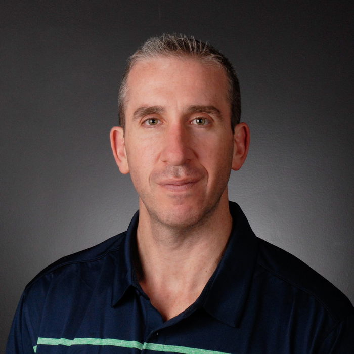 A man with short gray hair and a neutral expression wearing a dark polo shirt against a plain dark background.