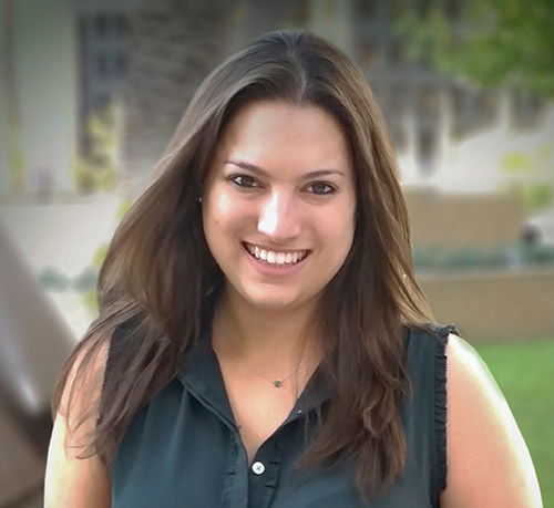 A woman with long brown hair smiles at the camera, standing outdoors.