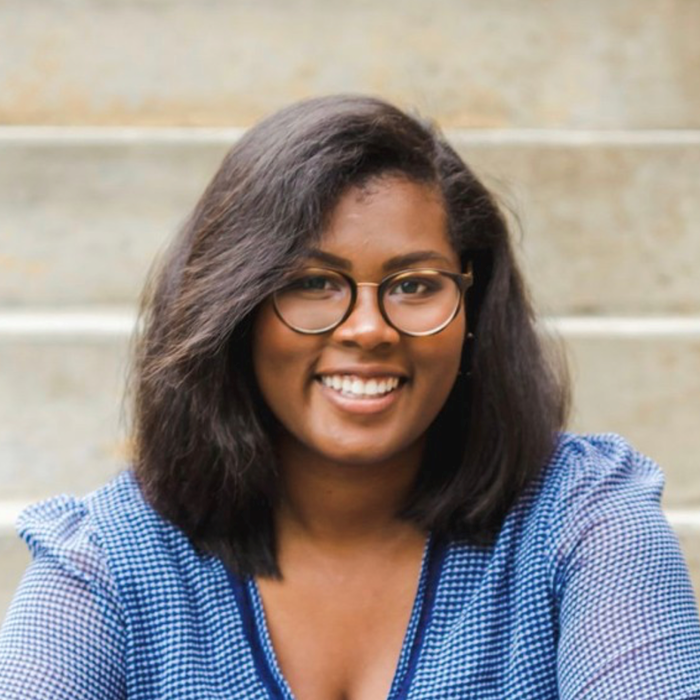 A person with shoulder-length hair and glasses smiles while sitting on steps, wearing a blue checkered top.