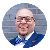 A person wearing glasses, a blue suit, and a bow tie smiles in front of a brick wall.