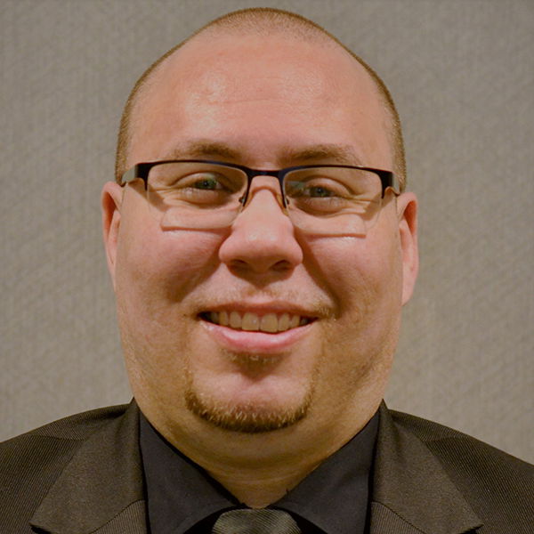 A man in glasses, wearing a black suit and tie, smiles at the camera against a plain background.