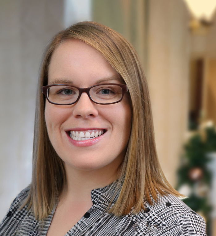A person with glasses and straight, shoulder-length hair smiles at the camera. They are wearing a patterned shirt.