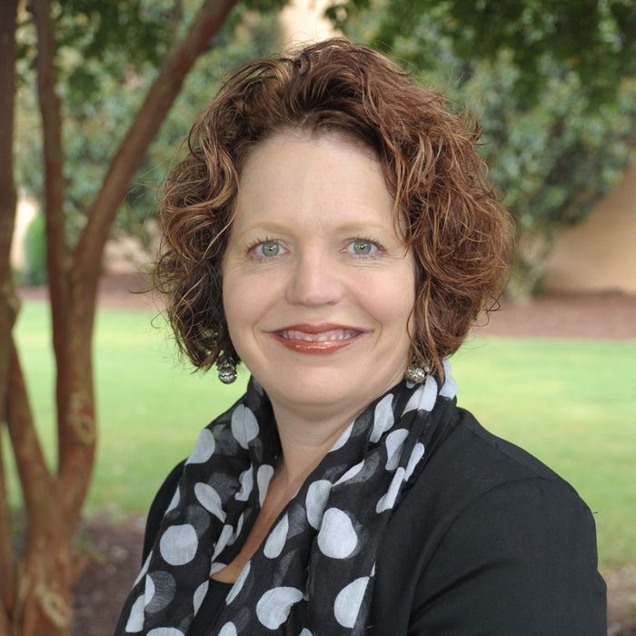 A woman with curly hair and a polka dot scarf smiles at the camera while sitting outdoors.