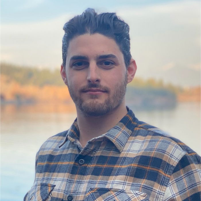 A person with a beard and plaid shirt stands in front of a serene lake and forest backdrop.