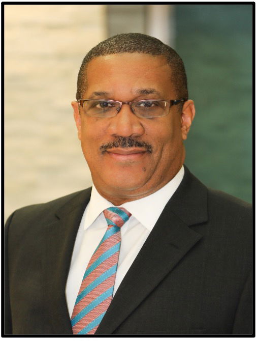 A man wearing glasses, a black suit, a white shirt, and a striped tie, looking directly at the camera with a neutral expression.