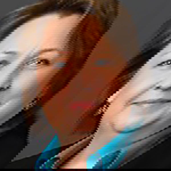 Portrait of a person with short, light brown hair, wearing a teal shirt and dark blazer, against a gray background.