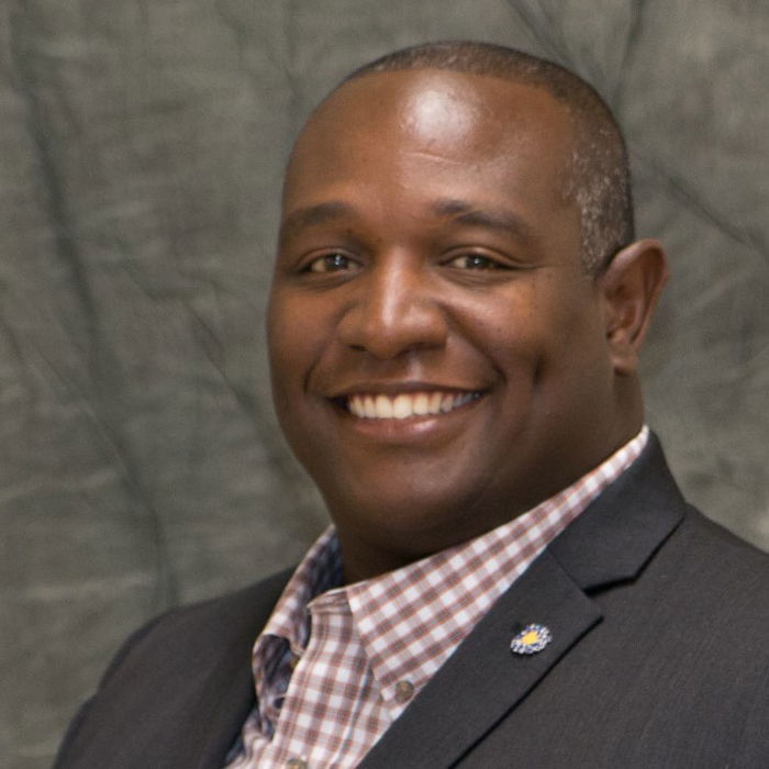 Smiling person wearing a checked shirt and dark blazer against a textured green background.