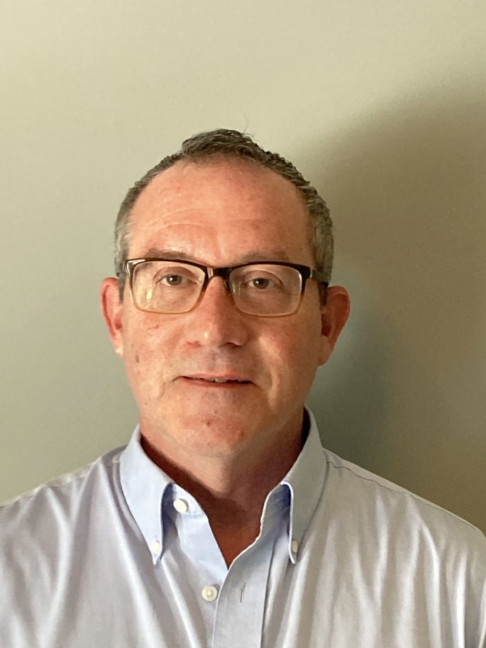 A man with glasses and short hair wearing a light blue collared shirt stands against a neutral background.