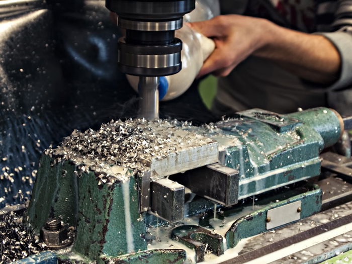 A person operates a milling machine, cutting a metal piece clamped in a vise, surrounded by metal shavings and coolant.