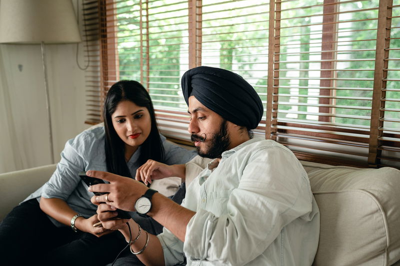 A man and a woman sit on a couch, drawn to the screen of a smartphone. The man, wearing a turban and watch, seems intent on the display. Sunlight filters through large windows with blinds in the room around them.
