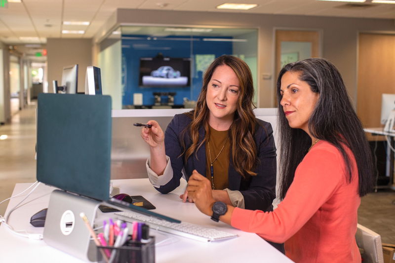 Two women sit at a desk in an office, studying a computer screen. One points at the digital drawing on the screen while the other listens attentively.