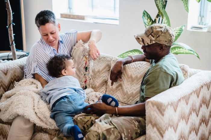 Two adults, one in a military uniform, and a child sit on a patterned sofa. The child is crawling as the adults smile and interact warmly—an everyday moment that human services nonprofits strive to support to BOOST family cohesion and well-being.