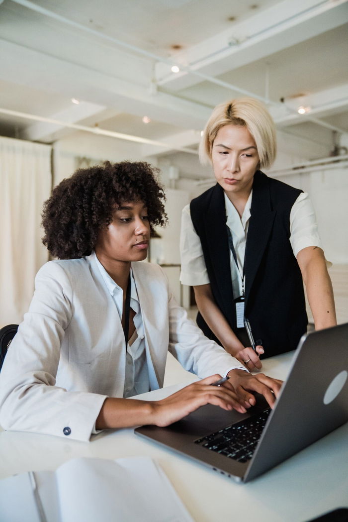 In a well-lit office space, two professionals collaborate at a table. One is using a laptop while the other points at the screen, both wearing smart business attire. They could be drafting new strategies for Michigan's Student Success Center to enhance credential programs.