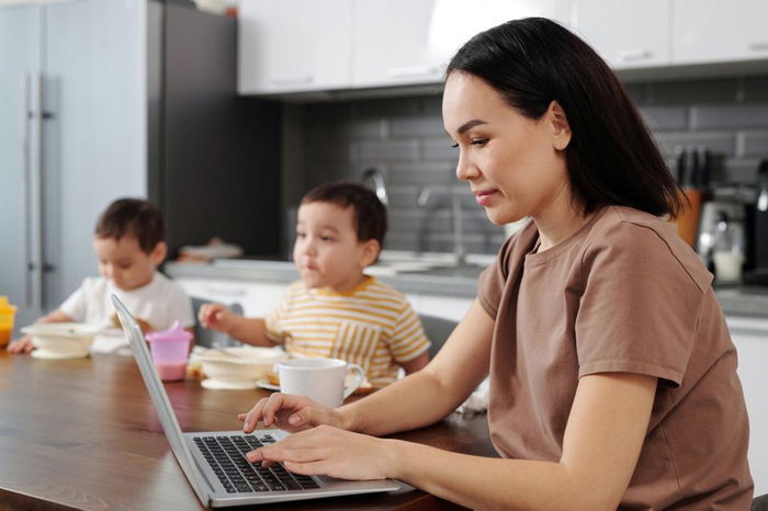 A woman types on a laptop at a kitchen table while two children eat in the background.