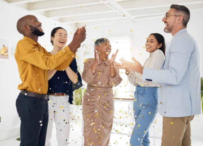 Five people in a room celebrate joyfully with confetti falling around them, smiling and clapping.