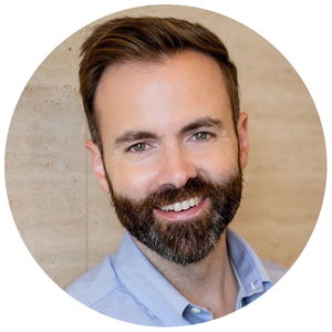 A bearded man in a light blue shirt smiles at the camera against a beige textured background.