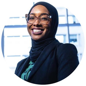 Person wearing glasses and a hijab, smiling, against a backdrop of a window with blurred buildings outside.