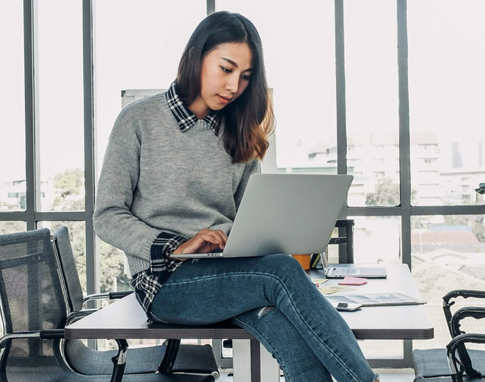 A person sits on a desk working on a laptop in a bright office setting, with large windows in the background.