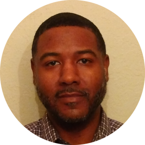 A person with a beard and short hair is looking directly at the camera. They are wearing a checkered shirt. The background is a plain, light-colored wall.