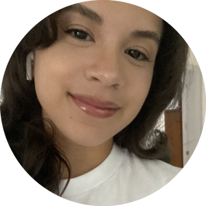 A person with long, wavy hair and wearing a white shirt smiles at the camera. They have earbuds in their ears.