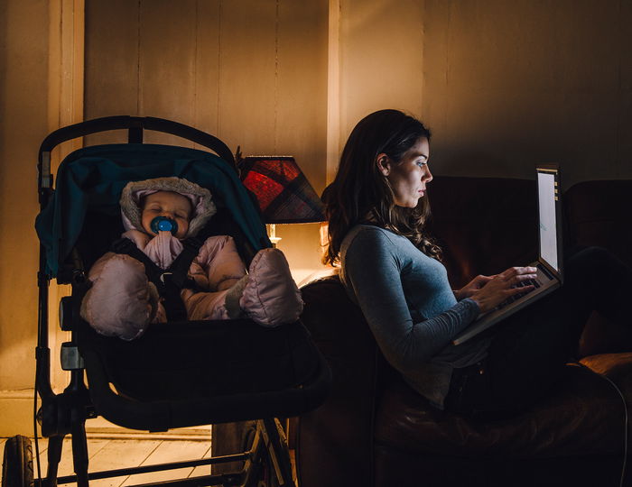 Woman working on a laptop sits on a couch next to a sleeping baby in a stroller.