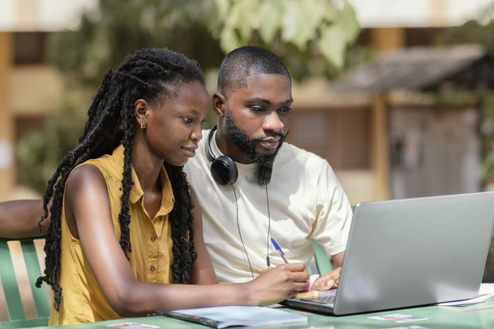 Two people sit together outdoors, focused on a laptop. Engrossed in their dual enrollment course, one takes notes with a pen while headphones rest around the man's neck.