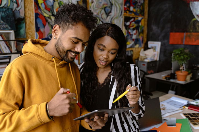 In an art studio filled with vibrant artwork and lush plants, two people engage in a direct assessment as they examine a tablet. With a red pencil in one hand and a yellow in the other, they collaborate creatively.